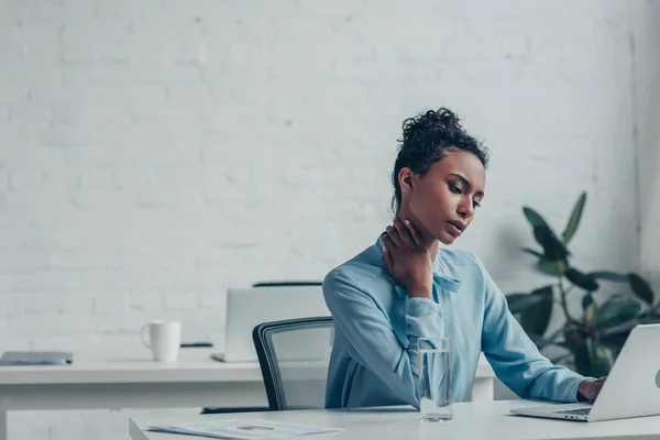 Hermosa mujer de negocios afroamericana que sufre de dolor de cuello mientras está sentada en el lugar de trabajo - foto de stock