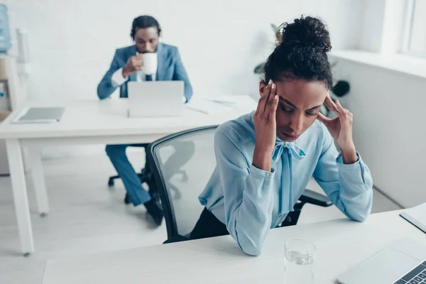 Enfoque selectivo de la mujer de negocios afroamericana que sufre de dolor de cabeza mientras está sentada en la oficina cerca de colega beber café - foto de stock