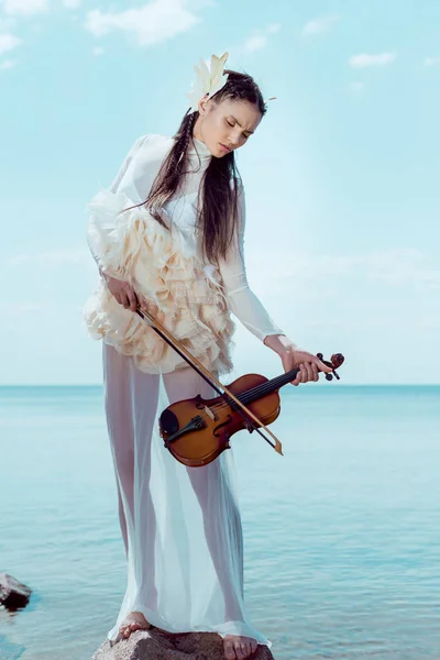 Tender woman in white swan costume with violin standing on blue river and sky background — Stock Photo