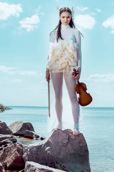 Baixo ângulo vista de mulher elegante em traje de cisne branco com violino em pé no fundo azul do rio e céu — Fotografia de Stock