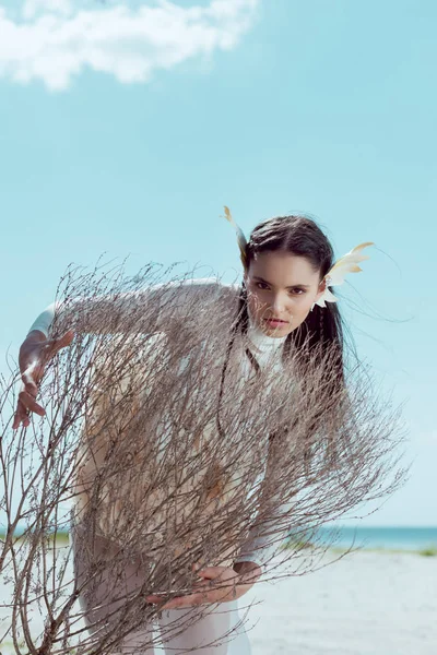 Mujer joven en traje de cisne blanco de pie detrás de un arbusto seco, mirando a la cámara - foto de stock
