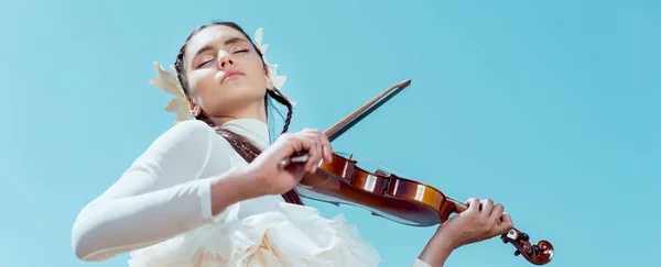 Vista de ángulo bajo de la tierna mujer en traje de cisne blanco de pie sobre fondo azul cielo con violín - foto de stock