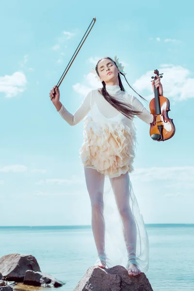 Belle femme en costume de cygne blanc debout sur fond de rivière avec violon — Photo de stock
