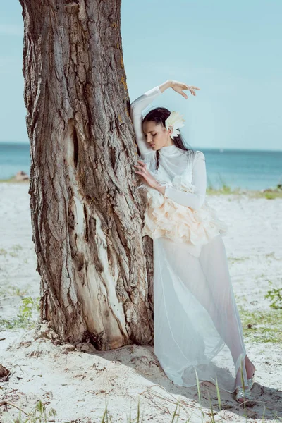 Mujer joven en traje de cisne blanco de pie cerca de tres troncos en la playa - foto de stock