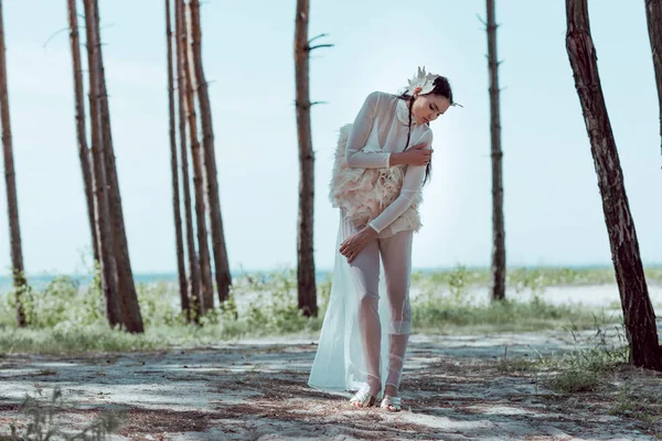 Beautiful woman in white swan costume standing on sandy beach near trees — Stock Photo