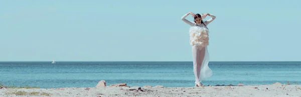 Панорамний вид на елегантну жінку в білому лебединому костюмі, що стоїть на піщаному пляжі — стокове фото