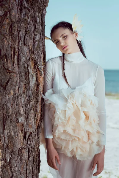 Elegante Frau im weißen Schwanenkostüm steht neben drei Baumstämmen und blickt in die Kamera — Stockfoto