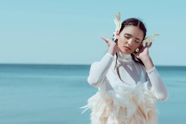 Tierna mujer en traje de cisne blanco de pie sobre el río azul y el fondo del cielo, sosteniendo la mano cerca de la cara - foto de stock