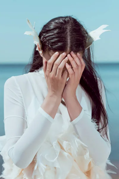 Upset woman in white swan costume standing on blue river and sky background, covering face by hands — Stock Photo