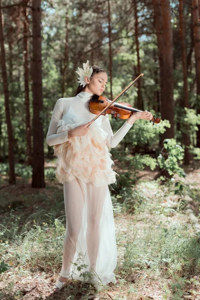 Elegante mujer en traje de cisne blanco de pie sobre el fondo del bosque, tocando el violín - foto de stock