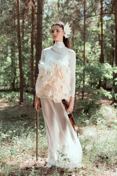 Mujer joven en traje de cisne blanco de pie sobre el fondo del bosque con violín, mirando hacia otro lado - foto de stock