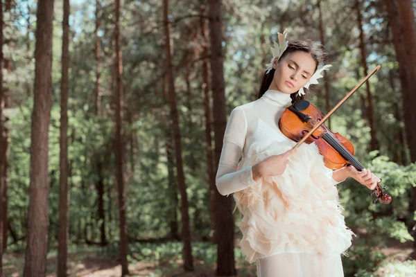 Femme rêveuse en costume de cygne blanc debout sur fond de forêt, jouant sur violon — Photo de stock