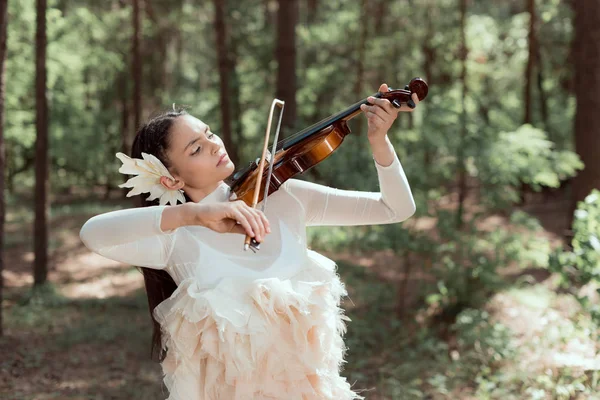 Mulher morena em traje de cisne branco em pé no fundo da floresta, tocando violino, olhando para longe — Fotografia de Stock