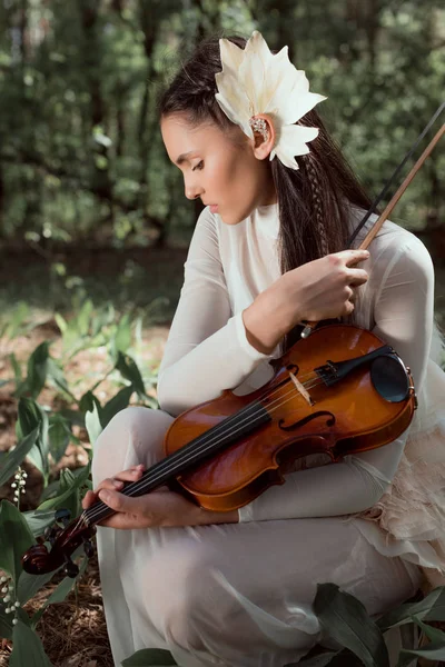 Mujer joven en traje de cisne blanco sentada en el suelo con el fondo del bosque, mirando hacia otro lado, sosteniendo violín - foto de stock