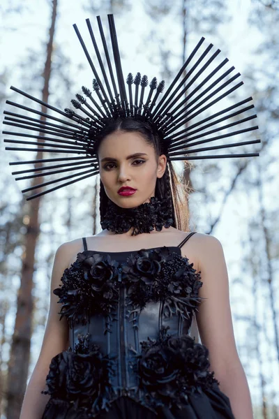 Baixo ângulo vista de mulher elegante em traje de bruxa em pé no fundo da floresta, olhando para a câmera — Fotografia de Stock