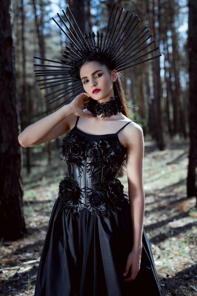 Enfoque selectivo de la mujer elegante en traje de bruja de pie sobre el fondo del bosque, sosteniendo la mano cerca del cuello, mirando a la cámara - foto de stock