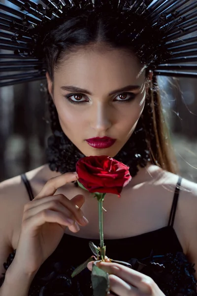 Retrato de mujer hermosa en traje de bruja mirando a la cámara, sosteniendo rosa roja en las manos — Stock Photo