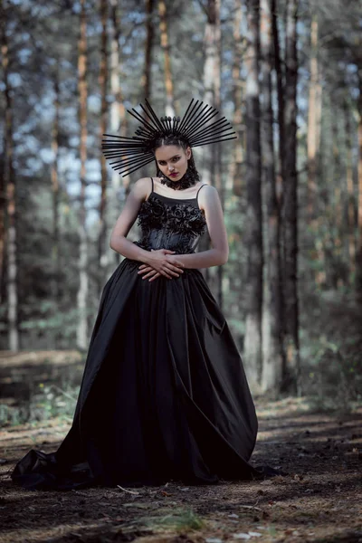 Mulher elegante em traje de bruxa em pé no fundo da floresta, olhando para a câmera — Fotografia de Stock