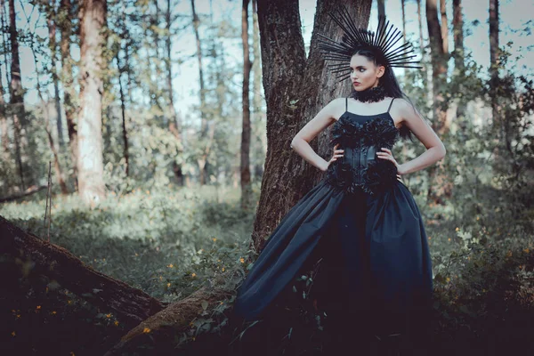 Femme élégante en costume de sorcière debout sur fond de forêt avec les mains sur les hanches, regardant loin — Photo de stock