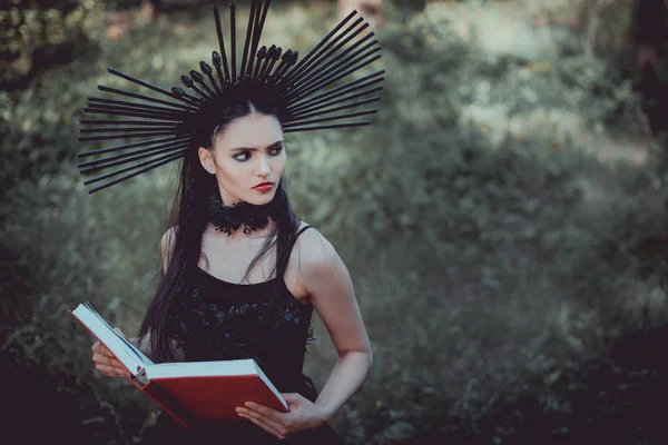 Vue grand angle de belle femme en costume de sorcière debout sur fond de forêt, tenant le livre rouge, regardant loin — Photo de stock