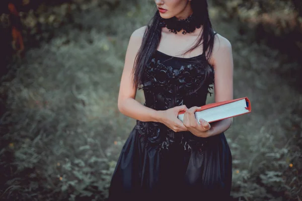 Vista recortada de la mujer en traje de bruja de pie sobre el fondo del bosque con libro rojo - foto de stock