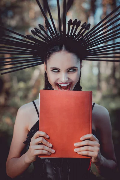 Mulher louca em traje de bruxa preta com coroa na cabeça grimacing, segurando livro vermelho — Fotografia de Stock