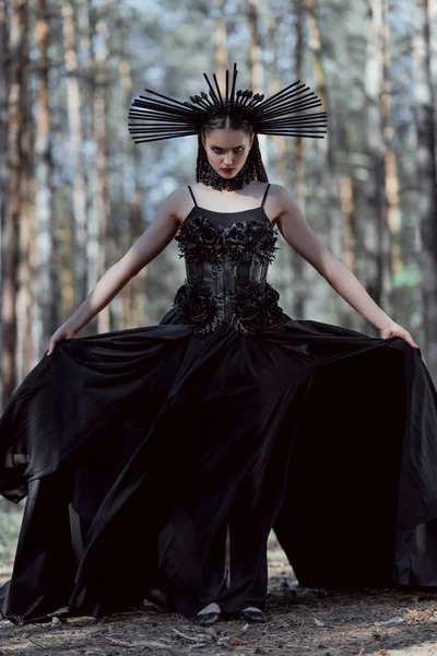 Mulher elegante segurando bainha de traje de bruxa, de pé no fundo da floresta, olhando para a câmera — Fotografia de Stock