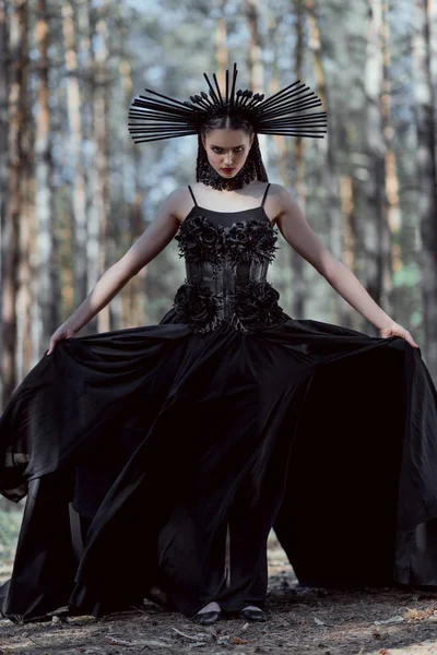 Elegant woman holding hem of witch costume, standing on forest background, looking at camera — Stock Photo