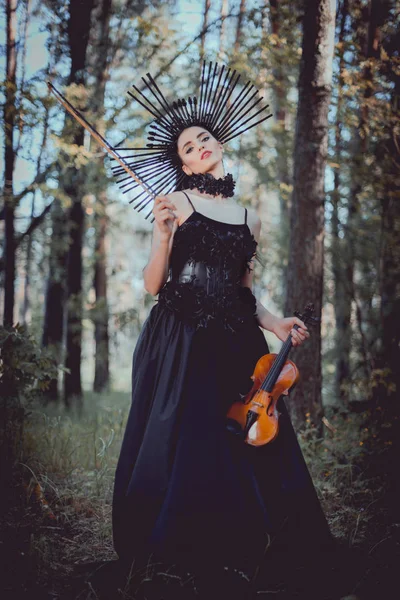 Low angle view of young woman in witch costume standing on forest background, holding violin — Stock Photo