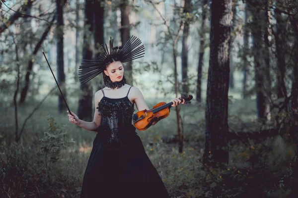 Mujer elegante en traje de bruja de pie sobre el fondo del bosque, mirando el violín - foto de stock