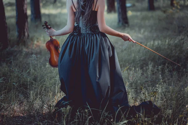 Back view of woman in witch costume walking on forest, holding violin — Stock Photo