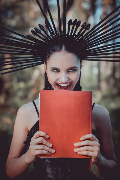 Сумасшедшая женщина в черном костюме ведьмы с короной на голове гримаса, держа красную книгу — стоковое фото
