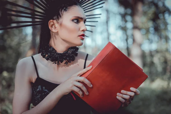 Красива жінка в костюмі відьми, що стоїть на лісовому фоні, тримає червону книгу, дивлячись — стокове фото