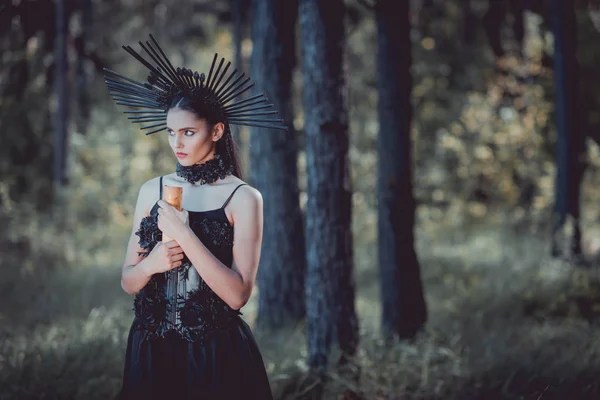 Belle femme en costume de sorcière debout sur fond de forêt, regardant loin, tenant défilement — Photo de stock