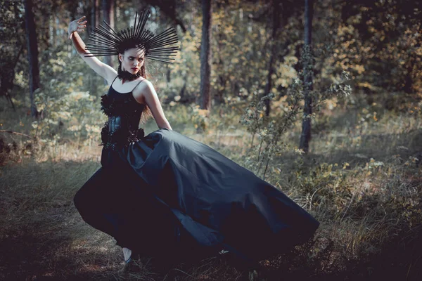 Mulher morena em traje de bruxa com coroa na cabeça em pé no fundo da floresta — Fotografia de Stock