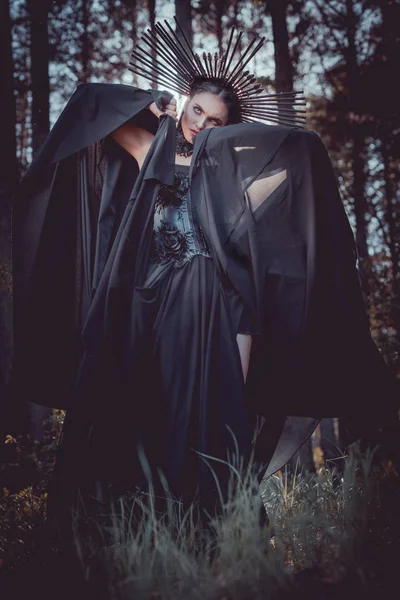 Femme brune en costume de sorcière et couronne sur la tête tenant la main avec du textile noir près du visage, détournant les yeux — Photo de stock