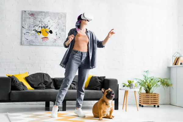 Дівчина з барвистим волоссям і гарнітурою VR, що стоїть біля собаки у вітальні — стокове фото