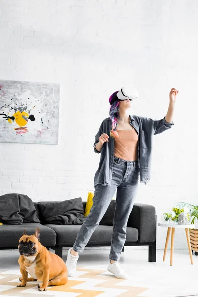 Chica con el pelo colorido y auriculares VR de pie cerca del perro y el gesto en la sala de estar - foto de stock