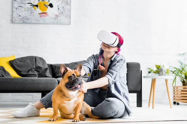 Girl with colorful hair and VR headset sitting on floor and petting dog in cosy room — Stock Photo