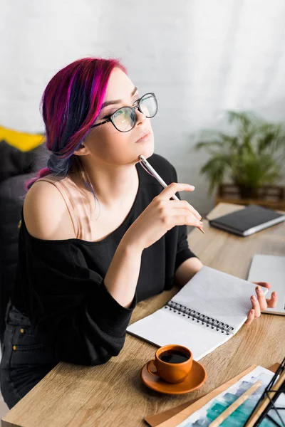 Belle fille avec des cheveux colorés assis derrière la table et regardant loin pensivement — Photo de stock