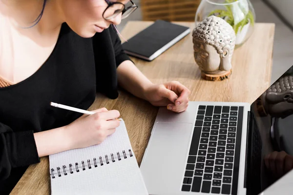 Schöne Mädchen sitzt hinter Tisch mit Laptop und schreibt in Notizbuch — Stockfoto