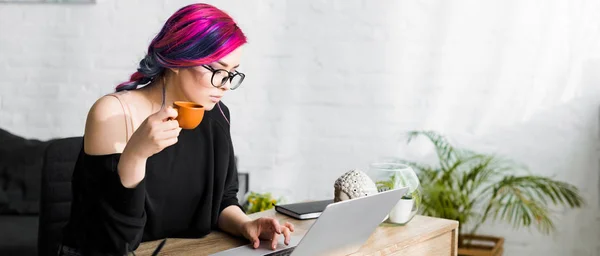 Plan panoramique de belle fille avec des cheveux colorés boire du café tout en étant assis à table et en utilisant un ordinateur portable — Photo de stock