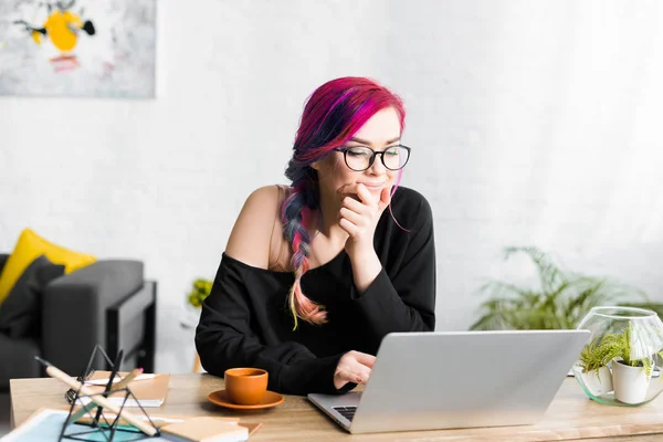 Chica hipster con el pelo colorido sentado detrás de la mesa y mirando el ordenador portátil pensativamente - foto de stock