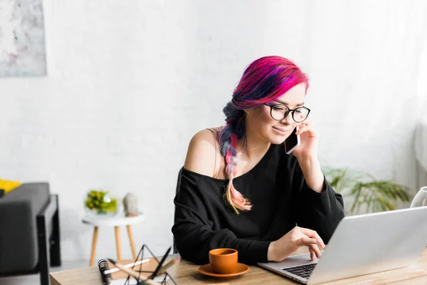 Chica hipster con el pelo colorido sentado detrás de la mesa, utilizando el ordenador portátil y hablando en el teléfono inteligente - foto de stock