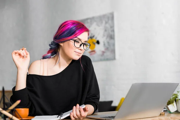 Chica con el pelo colorido sentado en el escritorio y tomando notas en la sala de estar - foto de stock