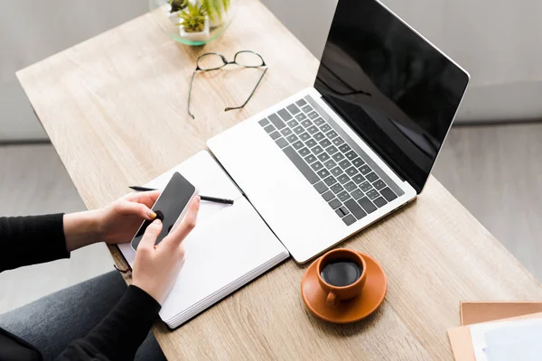 Cropped view of woman using smartphone and sitting behind table with laptop, notebook and cup of coffee — Stock Photo