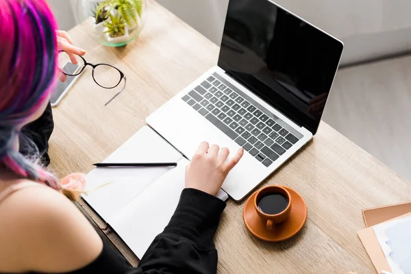 Cropped view of woman using laptop while sitting at table with notebook and cup of coffee — Stock Photo