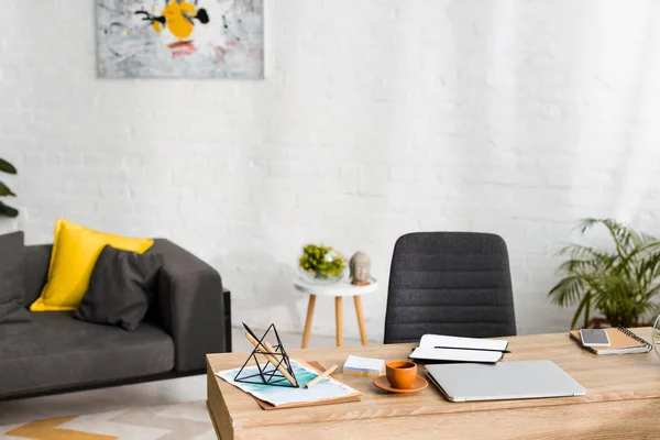 Mesa de madera con cuadernos, portátil y taza de café en el salón con sofá - foto de stock