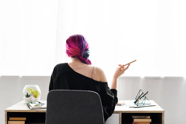 Back view of hipster girl holding pencil and sitting at table in living room — Stock Photo