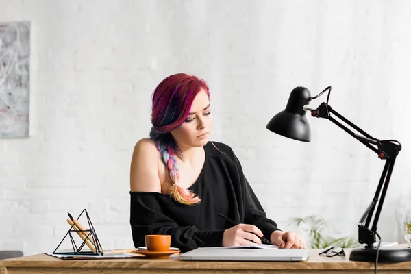 Atractiva chica hipster escribiendo algo mientras está sentado detrás de la mesa con el ordenador portátil, taza de café y lámpara - foto de stock